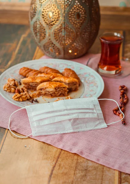 Turkish sweets baklava, kalburabasti and tea on ramadan 2020 on a plate and next a protective medical mask and decorative patterned vase.  Photos with shallow depth of field. Focus on the mask