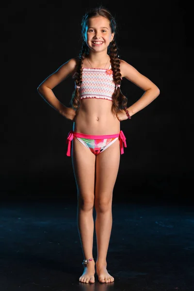 穿泳装的年轻少女赤脚站在黑色的背景上 漂亮的孩子 深色的头发和美丽的脸 Adorably 比基尼细长青春期 — 图库照片