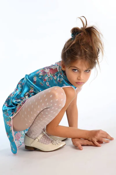 Beautiful girl in an asian blue dress is sitting on a white background. Elegant attractive child with a slender body and long legs in white pantyhose. The young model 9 years old in fashion style. — Stock Image Beautiful Girl Asian Blue Dress Sitting White Background Elegant Attractive Stock Image