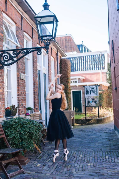 Young ballerina woman with red hair in ballet costume and pointe shoes is in beautiful pose dancing on street. Ballerina is posing in the streets. Girl dancing outdoors. Street ballet and performance