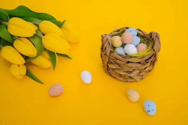 Easter holiday decorations on yellow background. Colored painting speckled eggs in basket. Creative spring composition with Easter elements - flowers and eggs. Celebrating Easter concept at spring.