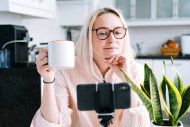 Mutfakta oturan mutlu bir kız ve elinde video çağrısı var. Genç bir kadın, arkadaşıyla ya da ailesiyle görüntülü konuşma yapmak için akıllı telefonu kullanıyor. Vlogger webinar kaydediyor. Kadın kameraya bakıyor ve el sallıyor.. 