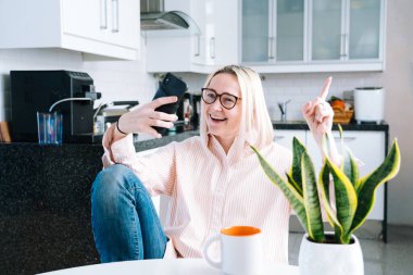 Mutfakta oturan mutlu bir kız ve elinde video çağrısı var. Genç bir kadın, arkadaşıyla ya da ailesiyle görüntülü konuşma yapmak için akıllı telefonu kullanıyor. Vlogger webinar kaydediyor. Kadın kameraya bakıyor ve el sallıyor.. 