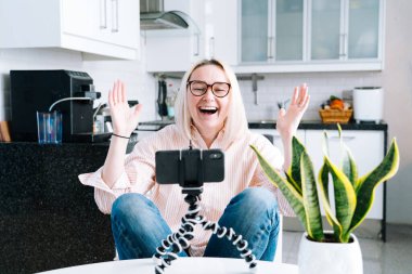 Mutfakta oturan mutlu bir kız ve elinde video çağrısı var. Genç bir kadın, arkadaşıyla ya da ailesiyle görüntülü konuşma yapmak için akıllı telefonu kullanıyor. Vlogger webinar kaydediyor. Kadın kameraya bakıyor ve el sallıyor.. 