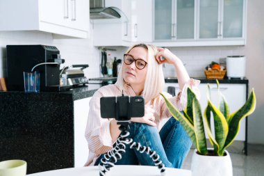 Mutfakta oturan mutlu bir kız ve elinde video çağrısı var. Genç bir kadın, arkadaşıyla ya da ailesiyle görüntülü konuşma yapmak için akıllı telefonu kullanıyor. Vlogger webinar kaydediyor. Kadın kameraya bakıyor ve el sallıyor.. 