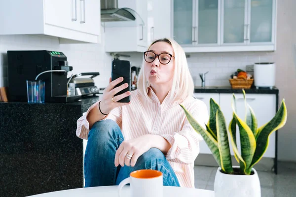 Happy girl sitting at home kitchen and holding videocall. Young woman using smartphone for video call with friend or family. Vlogger recording webinar. Woman looking camera and waving greeting hands.