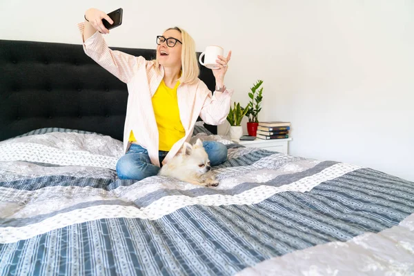 Woman using smartphone for video call with friend or family. Girl blogger with dog siting at bedroom and talking with followers. Vlogger recording webinar. Person sit on bed and looking to smartphone