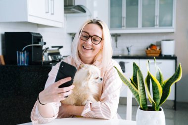 Mutfakta oturan mutlu bir kız ve elinde video çağrısı var. Genç bir kadın, arkadaşıyla ya da ailesiyle görüntülü konuşma yapmak için akıllı telefonu kullanıyor. Vlogger webinar kaydediyor. Kadın kameraya bakıyor ve el sallıyor.. 