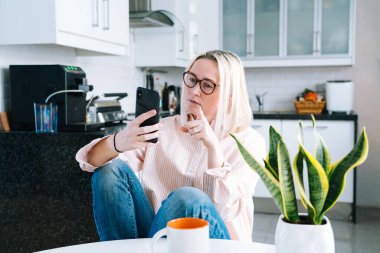 Mutfakta oturan mutlu bir kız ve elinde video çağrısı var. Genç bir kadın, arkadaşıyla ya da ailesiyle görüntülü konuşma yapmak için akıllı telefonu kullanıyor. Vlogger webinar kaydediyor. Kadın kameraya bakıyor ve el sallıyor.. 