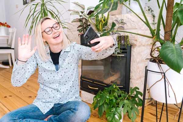 Happy girl sitting at living room and holding videocall. Young woman using smartphone for video call with friend or family. Vlogger recording webinar. Woman looking camera and waving greeting hands.