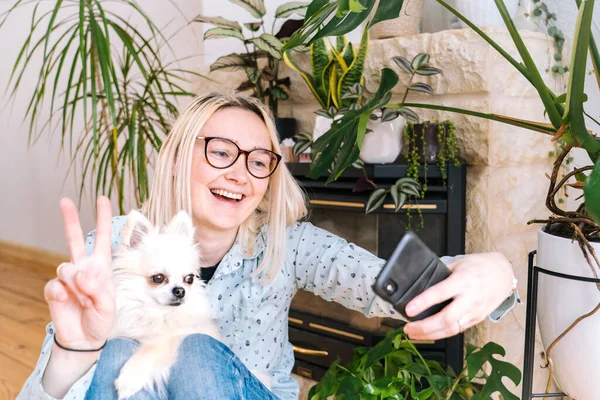 Happy girl sitting at home kitchen and holding videocall. Young woman using smartphone for video call with friend or family. Vlogger recording webinar. Woman looking camera and waving greeting hands