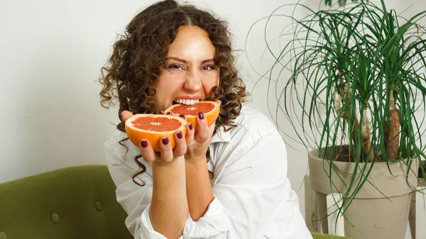 Красивая женщина кусает грейпфрут. Женщина с вьющимися волосами с грейпфрутом дома — стоковое фото