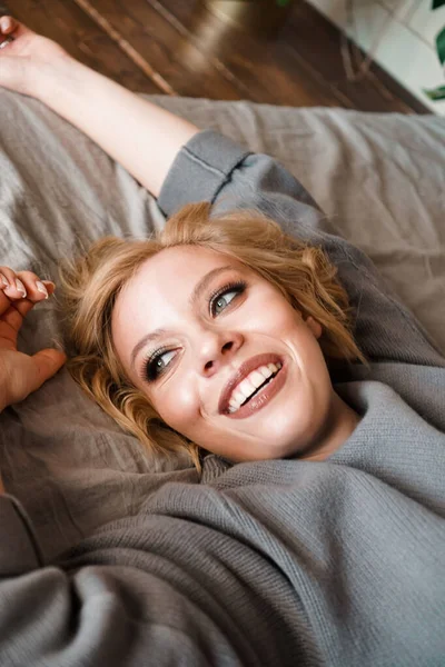 Красивая молодая женщина в сером свитере смеется лежа в постели — стоковое фото