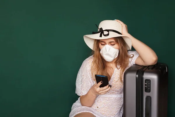 Chica turista en una máscara médica se sienta con el equipaje sobre un fondo verde —  Fotos de Stock