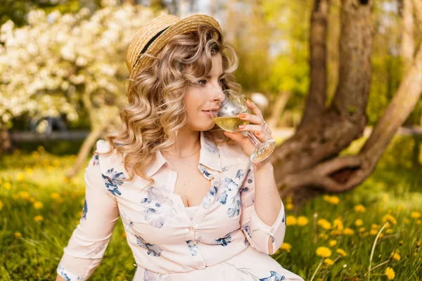 Bella giovane donna con i capelli biondi in cappello di paglia beve vino in giardino — Foto Stock