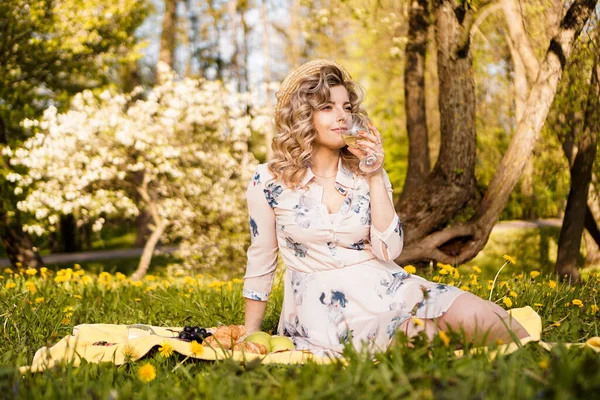Schöne junge Frau mit blonden Haaren im Strohhut trinkt Wein im Garten — Stockfoto