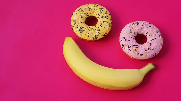 Två munkar och en banan ligger på rosa yta, bildar ett leende känslor — Stockfoto
