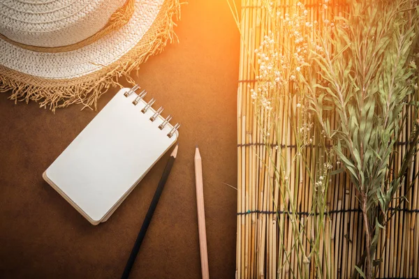 旅行概念以藤自然帽子与干燥花和树与空白页笔记本和铅笔在褐色皮革背景与轻的火光作用 — 图库照片