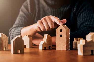 Ev tasarımı karar verme konsepti. İnsan eliyle seçtiği ev şeklindeki ahşap model kahverengi masa üzerinde.