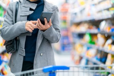 Yakın plan Asyalı kadın el bilgisayarı. Akıllı telefon kontrol fiyatları süpermarket fiyatlarıyla market alışveriş kartı ve sıra sıra ürünler karşılaştırılıyor.