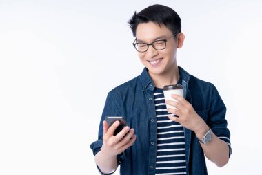 Zeki, çekici Asyalı gözlüklü erkek sıcak içecek, kahve el araması ve ferahlık ve neşeli günlük elbise beyaz arka plan ile akıllı telefon ekranına bakın.