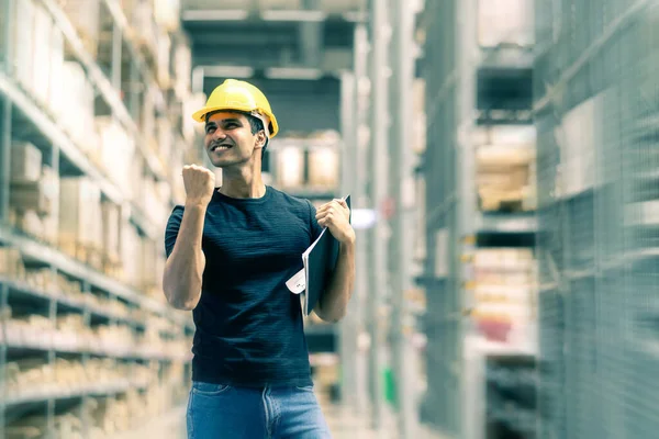 Smart Indian engineer man wearing safety helmet doing stock tick check and cardboard stock product management in factory warehouse background