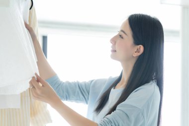 Güzel Asyalı ev hanımı sabah ışığı eşliğinde yürüme dolabında elbise seçip düzenler.