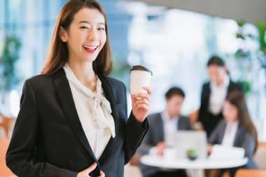 Zeki, çekici, Asyalı iş kadını, gülümseyen el, modern ofis geçmişine sahip sıcak bir içecek.