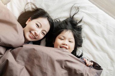 Mutlu Asyalı aile anı. Anne ve kız, beyaz yatak üzerinde battaniyeyle oynayıp uzanıyorlar.