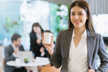 Zeki, çekici, Asyalı iş kadını, gülümseyen el, modern ofis geçmişine sahip sıcak bir içecek.