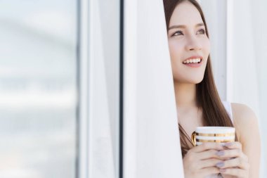 Güzel, çekici Asyalı beyaz kız tişörtü hafta sonu tatilinin tadını çıkar. İçi beyaz oda arka planında sıcak bir içecek ile birlikte.