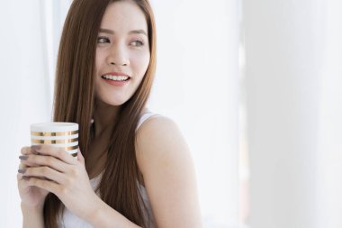 Güzel, çekici Asyalı beyaz kız tişörtü hafta sonu tatilinin tadını çıkar. İçi beyaz oda arka planında sıcak bir içecek ile birlikte.