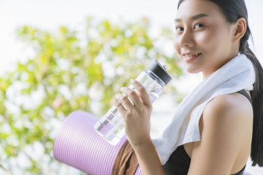 Güzel Asyalı kadın eli yoga minderi ve su şişesi tutuyor. Sağlıklı spor için yoga yapıyor. Bahçe geçmişi var.