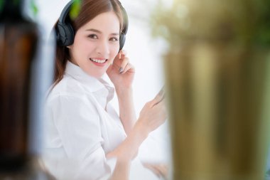 Güzel Asyalı beyaz elbiseli, uzun saçlı, kulaklık takıyor ve beyaz yataklı, beyaz ve neşeli beyaz yatak odası arka planlı akıllı bir telefon kullanıyor.