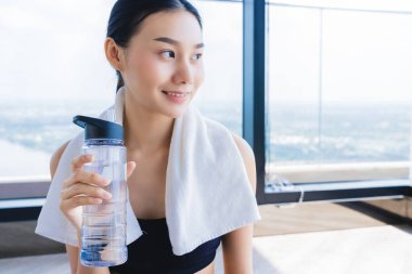 Spor yaptıktan sonra spor salonunda dinlenen Asyalı sağlıklı kadın saf su içeceği ve beyaz havlu tutuyor.