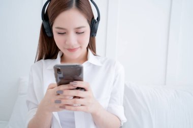 Güzel Asyalı beyaz elbiseli, uzun saçlı, kulaklık takıyor ve beyaz yataklı, beyaz ve neşeli beyaz yatak odası arka planlı akıllı bir telefon kullanıyor.