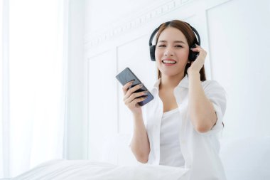 Güzel, çekici Asyalı kadın. Kulaklıktan şarkı dinle. Akıllı telefon, beyaz yatak odası, Asyalı uzun saçlı beyaz gömlekli kadın portresi.