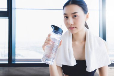Spor yaptıktan sonra spor salonunda dinlenen Asyalı sağlıklı kadın saf su içeceği ve beyaz havlu tutuyor.