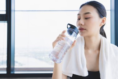 Spor yaptıktan sonra spor salonunda dinlenen Asyalı sağlıklı kadın saf su içeceği ve beyaz havlu tutuyor.