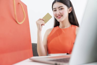 Orenge rengi elbiseli güzel Asyalı kadın dizüstü bilgisayarla alışveriş yapmayı ve indirim sezonunda kredi kartıyla ödeme yapmayı seviyor.