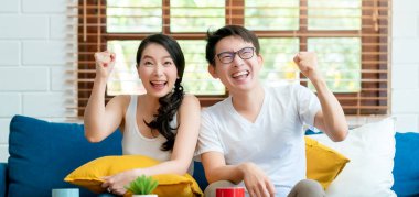 Asyalı evli çiftin el ele tutuşması ile başarıyı kutluyoruz. Kanepede birlikte spor maçı izliyoruz. Ev içi arka planda.