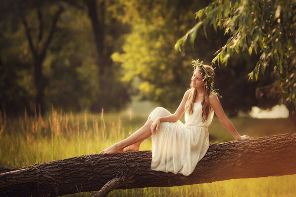 Young girl in a white dress is sitting on a tree in a forest in the rays of sunset light