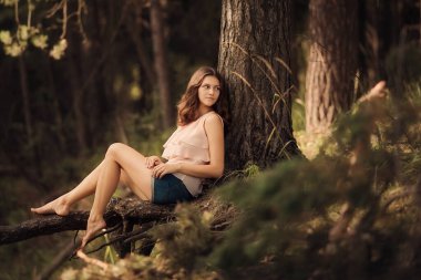 Güzel genç kız yaz ormanlarında ağaç köklerinde oturuyor.