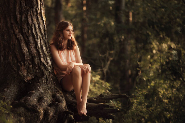 Beautiful young girl is sitting on tree roots in summer forest