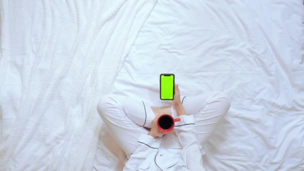 Выше вид женщины, держащей красную чашку и мобильный сидит в спальне — стоковое видео