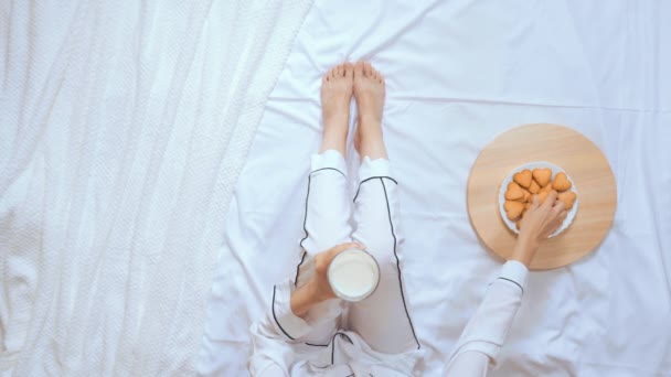 Weibchen im weißen Schlafanzug trinken Milch und essen Kekse im Bett — Stockvideo