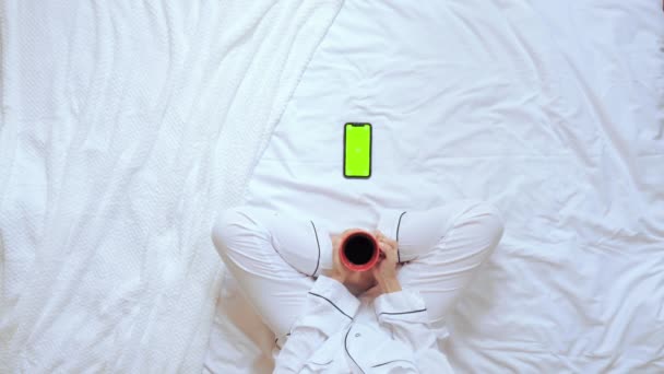 Vista sopra femmina tenendo tazza rossa guardando sul display mobile si siede in camera da letto — Video Stock