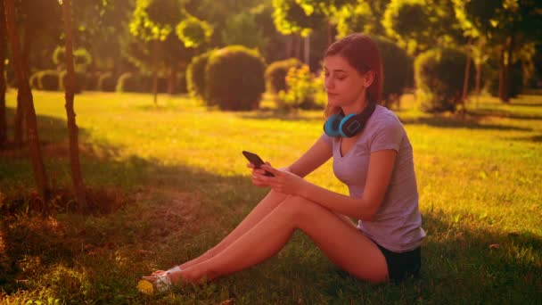 Cheerful female listening music sits on the green grass — Stock Video