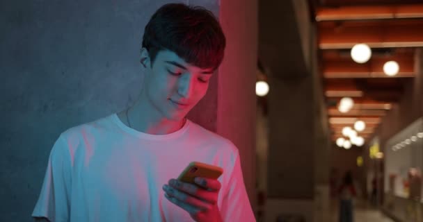 Joven Millenial Guy en Camiseta Blanca Usando su Smartphone, Sonriendo y Diciendo Wow mientras De Pie Inclinado a la Pared en Futuristic Neon Club Luces de Fondo. Concepto de comunicación . — Vídeos de Stock