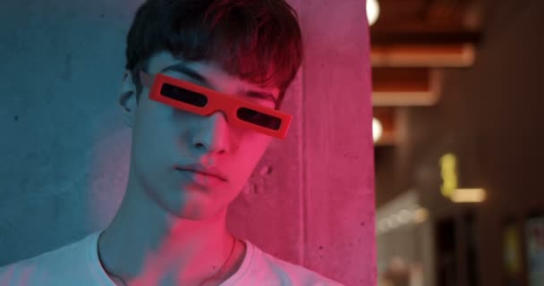Portrait of Cool Millenial Guy in Funny Party Glasses Standing Leaned his Head to Wall and Looking Straight to Camera at Futuristic Neon Club Lights (en inglés). Primer plano . — Vídeo de stock